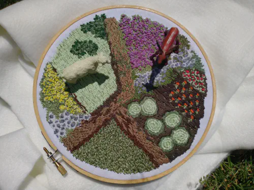 an embroidery hoop depicting a garden with a sheep and deer